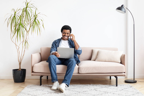 Rahatlamış Afro-Amerikalı adam evde dizüstü bilgisayar ve telefonla birden fazla iş yapıyor, iş hayatı dengesini belirtiyor. - Fotoğraf, Görsel