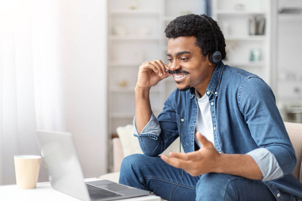 Ένας χαρούμενος νεαρός μαύρος με ακουστικά ασχολείται με βιντεοκλήση ή απομακρυσμένη εργασία στο λάπτοπ του σε ένα φωτεινό δωμάτιο. - Φωτογραφία, εικόνα