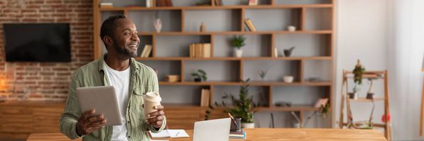 Смеющийся взрослый черный мужчина с планшетом и чашкой кофе наблюдает за свободным пространством в интерьере домашнего офиса. Работа и бизнес, анализ данных с помощью современных технологий, перерыв и отдых с новым приложением - Фото, изображение