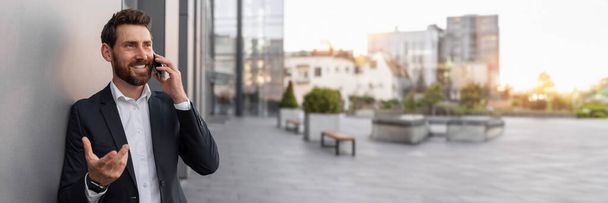 Vrolijke jonge Europese man baas met baard in pak gesprekken per telefoon, gesticulates in de buurt van kantoorgebouw op de stad straat. Communicatie op het werk, gesprek, financiën en opstarten, kopieerruimte - Foto, afbeelding