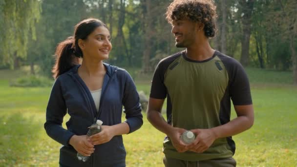 Two sport people smiling talking holding water bottles smile invitation join to exercise male Indian female Arabian friends couple man woman in park invite welcome workout fitness fit in city motivate - Footage, Video
