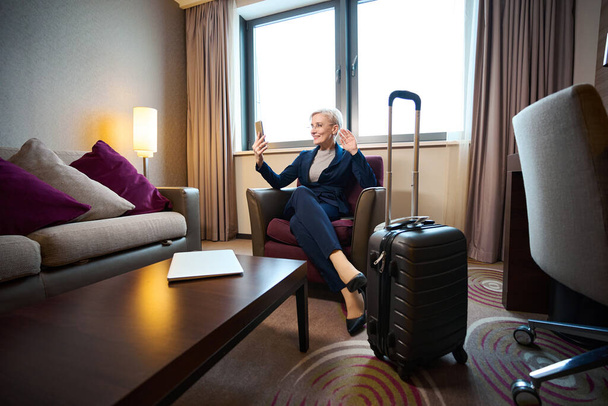 Volwassen blanke zakenvrouw zwaaiend met de hand tijdens videogesprek op smartphone in fauteuil in hotelkamer overdag. Concept van zakenreis, vakantie en reizen - Foto, afbeelding