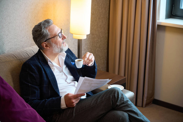 Keskittynyt aikuinen valkoihoinen liikemies, jolla on papereita, jotka juovat kahvia kupista ja katsovat muualle hotellihuoneessa päivällä. Työmatkan, loman ja matkustamisen käsite - Valokuva, kuva