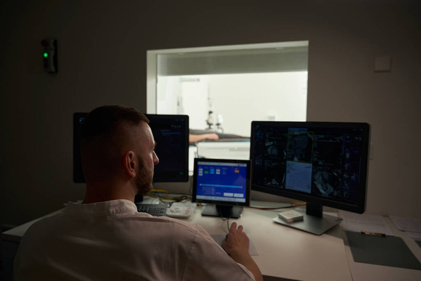 Gesundheitsexperte sitzt am Schreibtisch im Kontrollraum, während er CT-Scans auf dem Computermonitor betrachtet - Foto, Bild