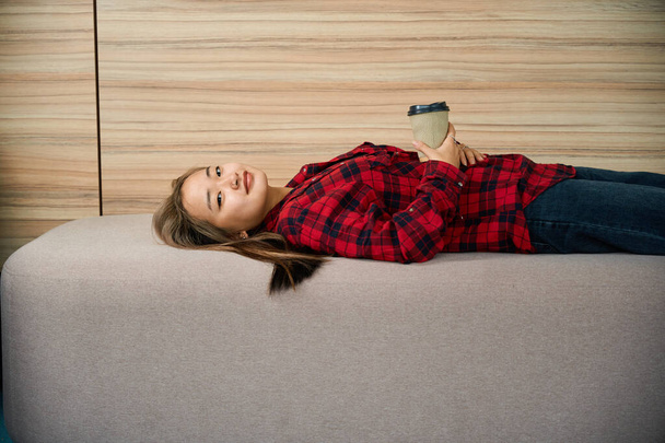Vista lateral de una joven empleada asiática seria con taza de café tumbada en el sofá y mirando a la cámara durante el descanso en la oficina de coworking. Concepto de trabajo independiente o remoto moderno - Foto, imagen