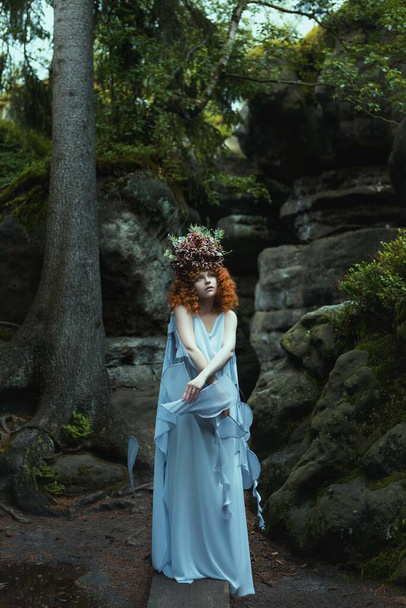 een vrouw in een blauwe jurk met een bloem op haar hoofd staat voor een rotswand. - Foto, afbeelding