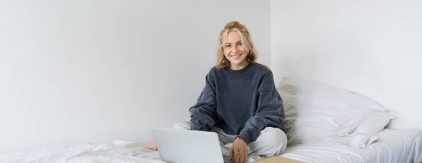 Retrato de una joven sonriente estudiando en su cama, trabajando desde casa en el dormitorio, sentada con portátil y cuadernos en pose de loto, luciendo feliz y relajada. - Foto, Imagen