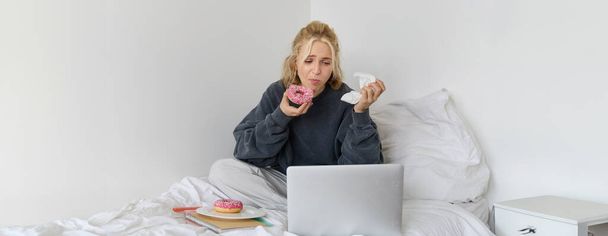 Portrait of woman watching sad show on laptop, eating doughnut and wiping tears with napkin, sitting on bed, spending time at home while being upset. - Photo, Image