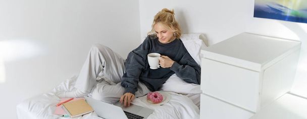 Πορτρέτο της νεαρής γυναίκας, φοιτήτρια που σπουδάζει στο κρεβάτι της, χαλαρώνοντας κατά την προετοιμασία της εργασίας, τρώγοντας ντόνατ, χρησιμοποιώντας φορητό υπολογιστή στο υπνοδωμάτιο και πίνοντας τσάι. - Φωτογραφία, εικόνα