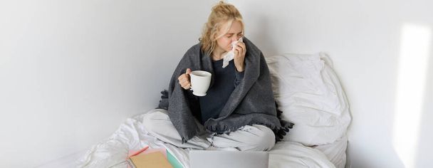 Ritratto di donna che prende l'influenza, starnutisce, si sente male, si siede sul letto con il computer portatile e lavora a distanza mentre si prende un raffreddore. - Foto, immagini