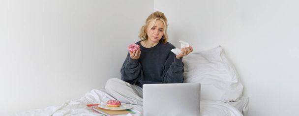 Portrait of sad, crying young woman, staying at home, sitting in bed with doughnut and comfort food, looking at something upsetting on laptop screen. - Photo, Image