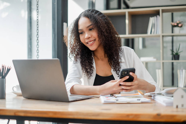 Uśmiechnięta młoda kobieta w białej marynarce siedzi przy drewnianym biurku, pracuje nad laptopem i trzyma smartfona. - Zdjęcie, obraz