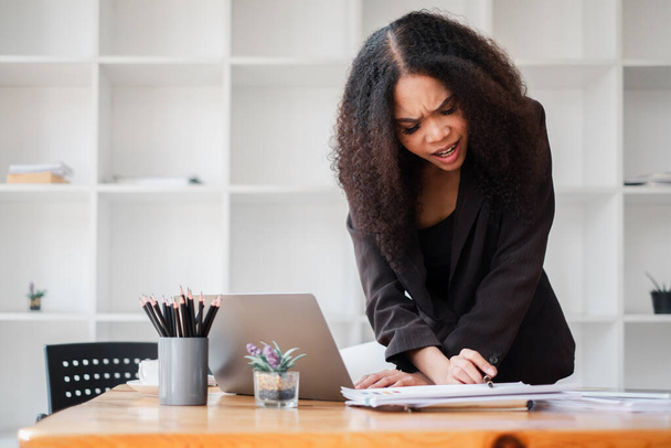 Mujer de negocios en una chaqueta oscura expresa preocupación al revisar documentos junto a una computadora portátil en su escritorio. - Foto, imagen