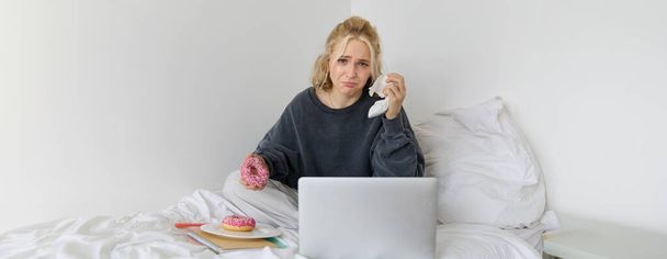 Portrét ženy, která se dívá na smutný film na notebooku, pláče a utírá si slzy, jí koblihy a pije čaj. - Fotografie, Obrázek