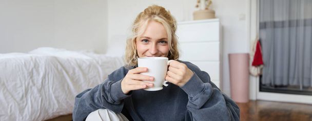 Obraz młodej nastolatki siedzącej w sypialni na podłodze, pijącej herbatę i cieszącej się dniem w domu, uśmiechniętej i patrzącej w kamerę. - Zdjęcie, obraz