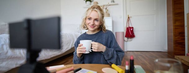 Portret van een leuke blonde vrouw, video content maker, vlogger opname op digitale camera in de slaapkamer, drinken van een kopje thee en praten met het publiek. - Foto, afbeelding