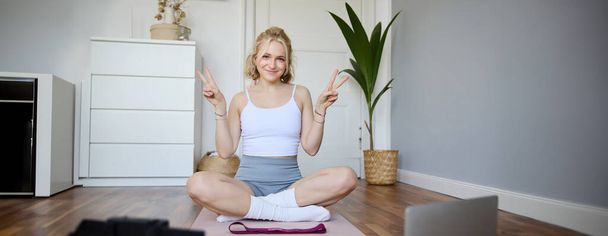 Портрет милої блондинки показує знак миру, посміхається, записує йогу вхід на цифрову камеру, тренується вдома на гумовому килимку в кімнаті. - Фото, зображення