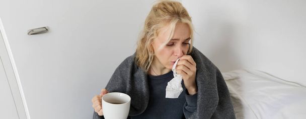Portret kobiety przeziębionej, chorującej w domu, pijącej herbatę, kichającej w serwetce, dmuchającej w nos, ma objawy. - Zdjęcie, obraz