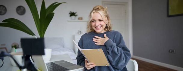 Portrait of smiling young cute woman, holding notebook, girl with laptop and planner in hands, sits in room, looks at digital camera, creates lifestyle vlog, talks to audience, reads script. - Photo, Image