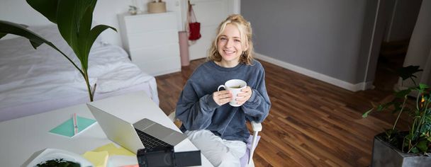 Imagem de jovem, influenciadora de mídia social, editando seu vídeo no laptop, senta-se em uma sala com computador e câmera digital, bebendo café, sorrindo para a câmera. - Foto, Imagem