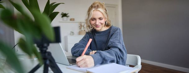 Portrait of young woman, lifestyle blogger, recording video of herself, making notes, writing in journal, sitting in front of laptop in a room and studying. - Photo, Image