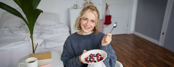 Retrato de una mujer rubia sonriente, desayunando, sosteniendo tazón y cuchara, sentada en el dormitorio, mirando feliz a la cámara. Concepto de estilo de vida - Foto, imagen