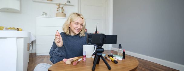 Retrato de mujer rubia sonriente registra un blog de estilo de vida, vlogger o artista de maquillaje grabando video para redes sociales, sosteniendo rímel, revisando productos de belleza para seguidores en línea. - Foto, imagen