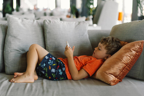 カーリーヘアの男の子は,電話でゲームに深く浸された枕付きの灰色のソファーで快適にリクライニングしています. 屋内でゆったりとした日々. - 写真・画像