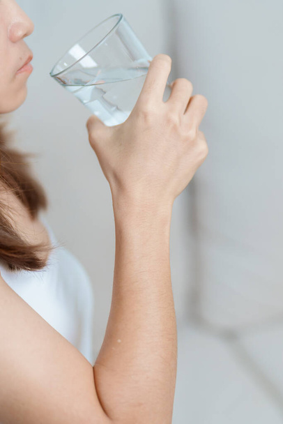 Ευτυχισμένη γυναίκα που κρατάει το ποτήρι με το νερό, γυναίκα που πίνει καθαρό νερό στον καναπέ στο σπίτι. Υγιεινός, αναζωογονητικός, τρόπος ζωής και καθημερινή έννοια ρουτίνας - Φωτογραφία, εικόνα