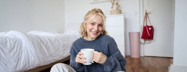 Portret van jonge mooie vrouw in casual kleding, zittend op de slaapkamervloer met kopje thee, drinken en glimlachen, kijkend naar de camera, het opnemen van een lifestyle vlog. - Foto, afbeelding
