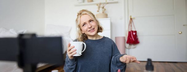 Porträt der blonden jungen Lifestylebloggerin, Frau nimmt Video auf, wie sie über Leben und Schönheit spricht, sitzt vor der Kamera, hält Tasse in der Hand, trinkt Tee, bloggt Inhalte für Social-Media-Account. - Foto, Bild