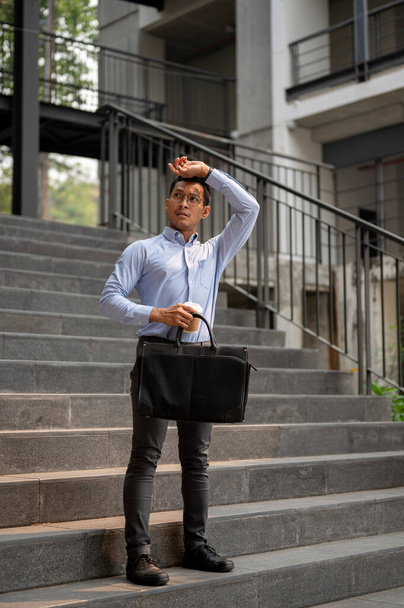 Egy kimerült ázsiai üzletember áll a lépcsőn, egy kávéscsészével és egy aktatáskával a kezében, fáradt arckifejezéssel törli a homlokát, meleget érez a naptól. üzletemberek koncepciója - Fotó, kép