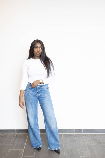 Eine junge Schwarze präsentiert einen zeitlosen, trendigen Look in einem taillierten weißen Oberteil gepaart mit klassisch blauen Jeans. Ihre Hand ruht auf ihrer Hüfte, eine goldene Uhr glänzt als Akzent. Sie steht gegen eine krasse - Foto, Bild