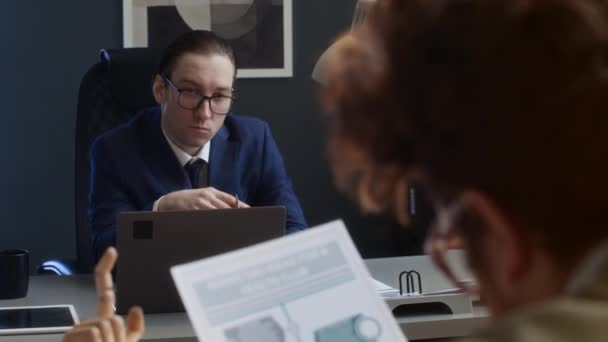 Zdjęcie przez ramię wkurzonego mężczyzny kaukaskiego menedżera korporacyjnego w okularach i garniturze siedzącego przy biurku w biurze, poprawiającego okulary, trzymającego się za ręce i patrzącego na rudego pracownika pracującego nad raportem - Materiał filmowy, wideo