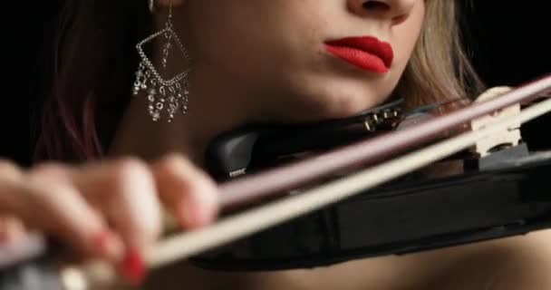 Capturée en vidéo, une casserole verticale du visage d'une violoniste à son torse la révèle jouant un violon électrique noir, orné d'une robe noire sans bretelles avec rouge à lèvres, ongles, boucles d'oreilles et cheveux blonds, sur fond noir - Séquence, vidéo