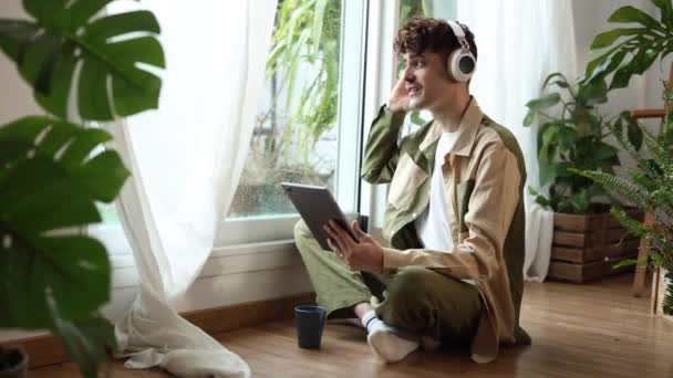 Ein nachdenklicher junger Mann sitzt am Fenster, gedankenverloren mit Musik und Tablet an einem gemütlichen, regnerischen Tag. - Filmmaterial, Video