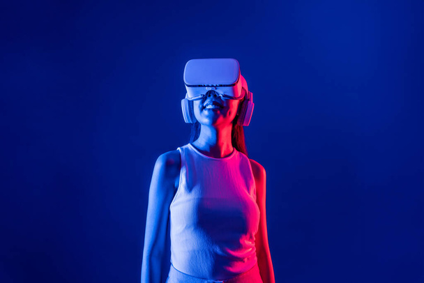 Smart female stand with surrounded by cyberpunk neon light wear VR headset connecting metaverse, futuristic cyberspace community technology. Elegant woman look generated virtual area. Hallucination. - Photo, Image