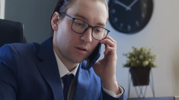 Střední detailní záběr narcistického pracovníka mužské kanceláře sedícího za stolem, který vede obchodní konverzaci na mobilním telefonu, dívá se na grafy, hladí si mastné vlasy a narovnává brýle - Záběry, video