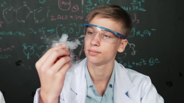 Zbliżenie chłopca sprawdzić roztwór chemiczny trzymając zlewkę na tablicy z teorii chemicznej. Inteligentny naukowiec robi eksperyment podczas analizy barwnego roztworu podczas noszenia fartucha laboratoryjnego. Budowanie - Materiał filmowy, wideo