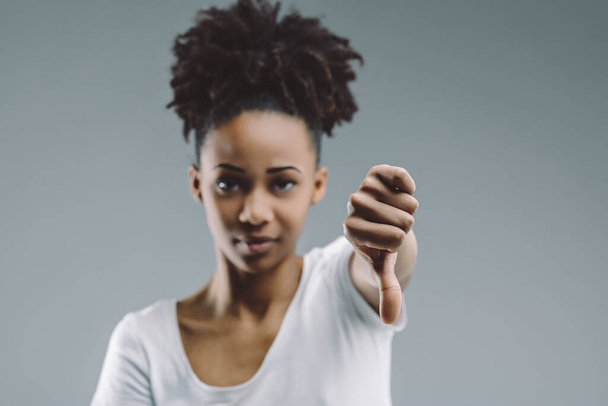Donna dai capelli afro dà un gesto pollice verso il basso, con uno sguardo esigente, impostato su uno sfondo semplice - Foto, immagini