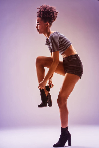 Jeune femme avant-gardiste en chemise rayée et short met sa botte, mettant en valeur la grâce et le style - Photo, image