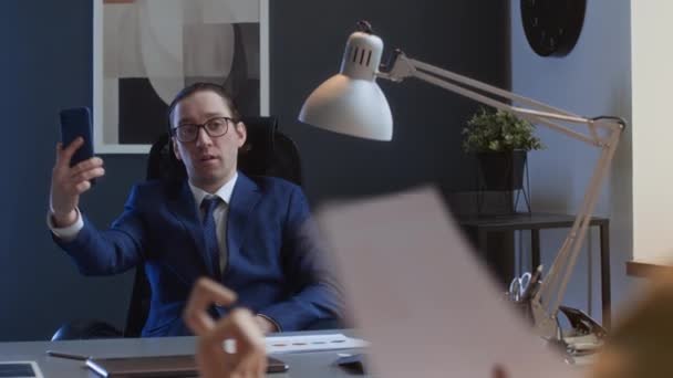 Medium over-shoulder shot of narcissistic Caucasian male corporate manager sitting at desk, preening, having video call, admiring himself, bullying and mocking subordinates work - Footage, Video