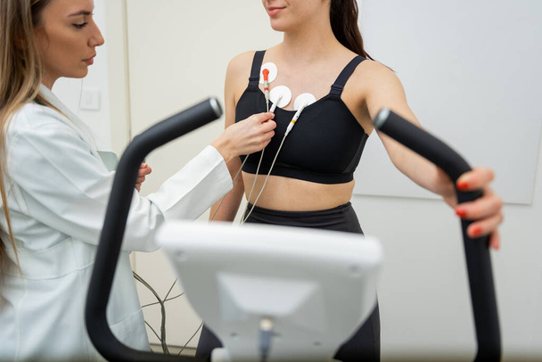Spor kıyafetleri içinde odaklanmış bir kadın egzersiz yaparken bir profesyonel göğsüne sensörler takıyor.. - Fotoğraf, Görsel