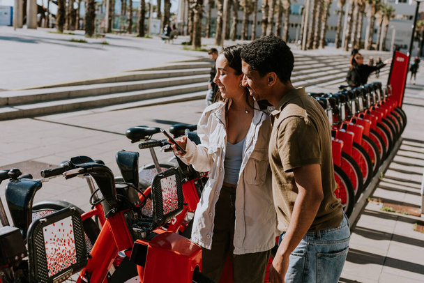 Giovane coppia che si gode una giornata ventilata scegliendo biciclette da una stazione di noleggio a Barcellona. - Foto, immagini