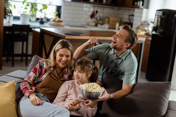 Trzyosobowa rodzina jest komfortowo położona na kanapie, ich twarze odzwierciedlają ekscytację i troskę, gdy dzielą się miską popcornu podczas napiętej nocy filmowej. - Zdjęcie, obraz