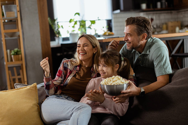 Kolmen hengen perhe lepää mukavasti sohvalla, heidän kasvonsa heijastavat jännitystä ja tarkkaavaisuutta, kun he jakavat popcornikulhon jännittävän elokuvaillan aikana. - Valokuva, kuva