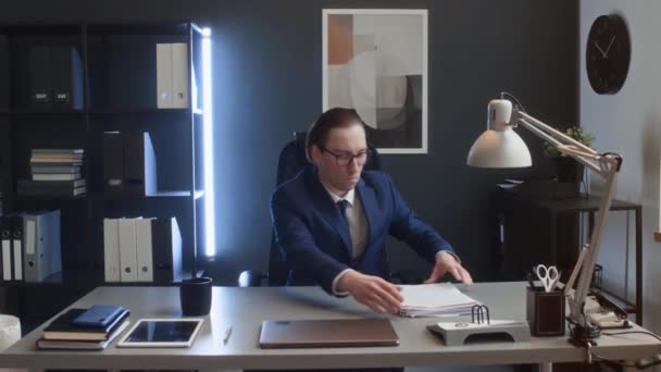 Medium volledige portret beelden van blanke mannelijke corporate werknemer in pak, bril zitten aan het bureau in het kantoor, rechttrekken stapels documenten, kijken naar de camera, glimlachen, poseren met geklemde handen - Video