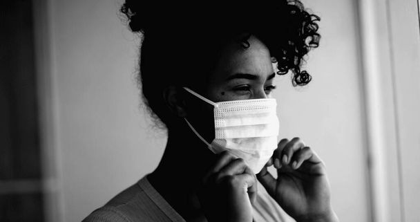 One young black woman putting covid face mask stuck at home in intense dramatic monochromatic portrait close-up face gazing at window from apartment - Photo, Image