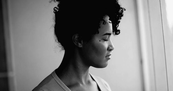 Jedna kontemplacyjna młoda czarna kobieta stojąca przy oknie patrząc w oddali z medytacyjnymi emocjami w dramatycznym monochromatycznym, czarno-białym. Zbliżenie profilu - Zdjęcie, obraz