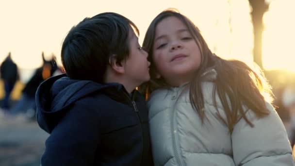 Momento de calentamiento entre el hermano pequeño y la hermana en el banco del parque compartiendo amor y besos en la mejilla durante el atardecer - Imágenes, Vídeo